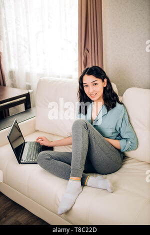 Bella femmina con sorriso positivo si siede nel divano vicino alla finestra utilizza generico computer laptop e connessione a internet ad alta velocità, rilassata a casa. Te moderno Foto Stock