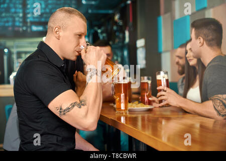 Vista dal lato del forte tatuato uomo seduto al tavolo e bere birra nei pub. Atletica Giovane maschio in camicia nera mantenendo la pinta con ale e godersi la serata con gli amici. Concetto di bevanda. Foto Stock