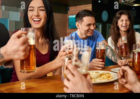 Felice compagnia ridendo e scherzando mentre si beve una gustosa birra nei pub. Allegro uomini e donne seduti insieme al grande tavolo, mangiare formaggio e salsicce, godendo di bevanda e di riposo. Concetto di felicità. Foto Stock