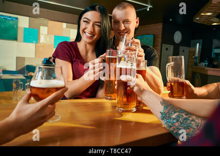 Vista frontale di allegro coppia seduta a tavola in bar e ridere con gli amici mentre si beve birra. I giovani in appoggio sul week-end con la società nel pub. Concetto di tempo libero e di felicità. Foto Stock