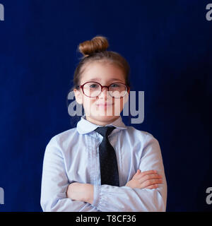 Un diligente studente della scuola in piedi con le braccia conserte. Fiducioso ragazza in età prescolare in uniforme con cravatta. Positivo il grado elementare pupilla in bicchieri su sfondo blu scuro. Carino felice schoolgirl Foto Stock