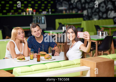 Allegro bella bruna mantenendo il telefono e tenendo selfie con gli amici nel bar. Funny Girl e ragazzo seduto di fronte a guardare fotocamera, sorridente e in posa mentre mangia hamburger gustosi. Foto Stock