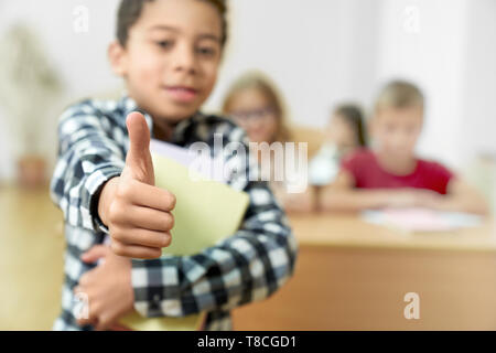 Stretta di mano maschio che mostra il pollice verso l'alto. Giovane bello schoolboy in piedi in aula, tenendo i libri. Gli alunni al desk in background. Foto Stock