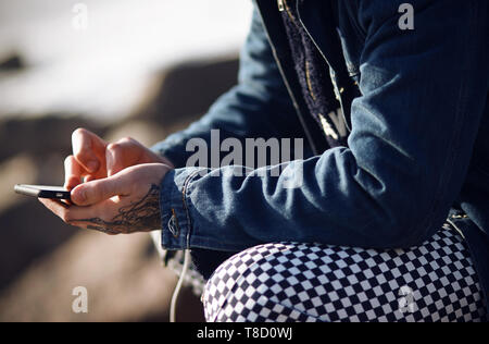 Un vestiti alla moda uomo in una camicia di denim e plaid pants detiene un toccare Telefono nella sua tatuato mani Foto Stock