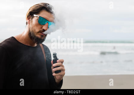 Uomo con barba e occhiali da sole di fumare siga elettronica all'aperto sulla spiaggia al tramonto. Giovani hipster vaping maschio sul fronte mare. Foto Stock