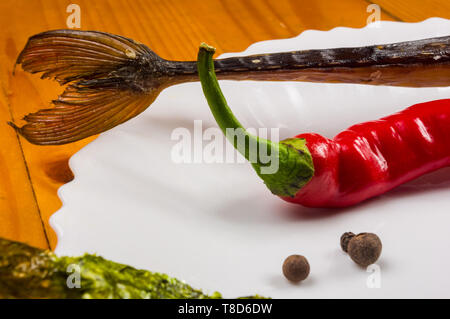 Still Life - aguglia affumicato con calce, basilico, cipollotti, peperoncino, nori chips, spezie, olio d'oliva in un bianco piatto di ceramica, su di un tavolo di legno Foto Stock