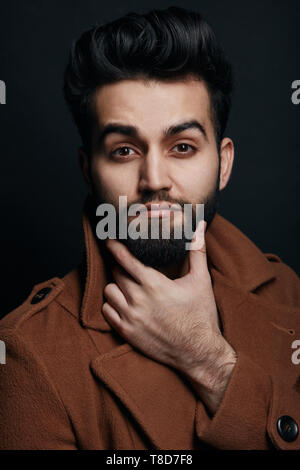 Barbuto uomo bello toccare la sua barba e guardando la telecamera. elegante macho rendendo la decisione, pianificazione il suo giorno, isolato sfondo nero, idea concept Foto Stock