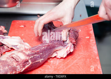 Closeup uomo caucasico chef macellaio mani di taglio del pezzo grezzo di filetto di maiale con coltello, su plastica tagliere professionale sul tavolo in metallo Foto Stock