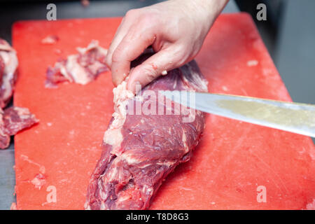 Closeup uomo caucasico chef macellaio mani di taglio del pezzo grezzo di filetto di maiale con coltello, su plastica tagliere professionale sul tavolo in metallo Foto Stock