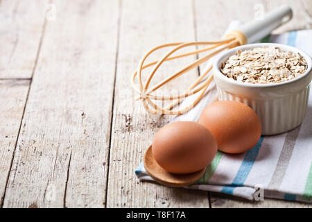 Fresco uova di pollo, fiocchi di avena nel recipiente di ceramica e arnese da cucina su tavola in legno rustico sfondo. Utensili da forno con copia spazio. Foto Stock