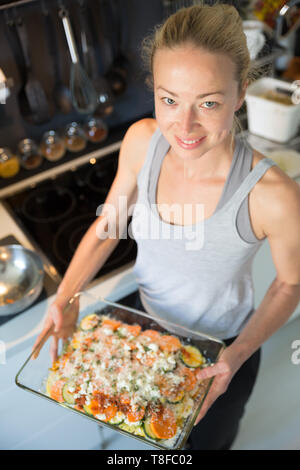 Sorridente giovane donna sana azienda e che mostra con orgoglio la cottura in vetro provare con fila piatto vegetariano ingredienti prima di metterla in forno. Foto Stock