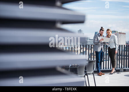 Due giovani uomini di affari che con la compressa in piedi su una terrazza fuori ufficio, funzionante. Foto Stock