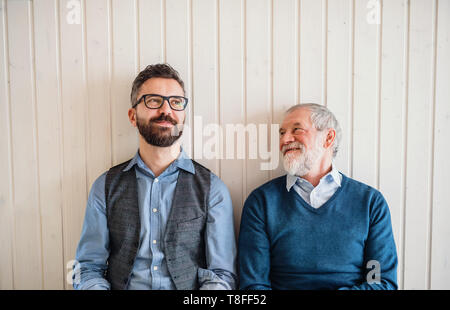 Un ritratto di adulto hipster figlio e padre senior seduto sul pavimento all'interno di home. Foto Stock