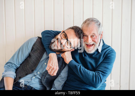 Un ritratto di adulto hipster figlio e padre senior seduto sul pavimento all'interno di home. Foto Stock