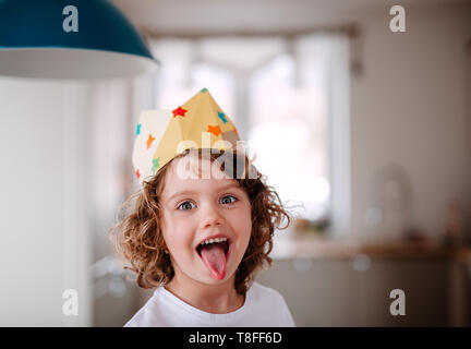 Una piccola ragazza con una corona di carta a casa, divertendosi guardando la telecamera. Foto Stock