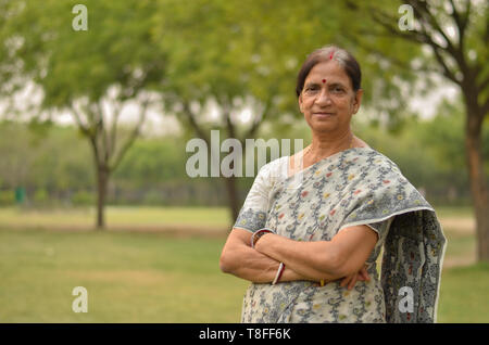 Donna anziana del nord intelligente in piedi, in posa per la macchina fotografica con le mani attraversate in un parco che indossa bianco / grigio saree in estate a Nuova Delhi, India Foto Stock