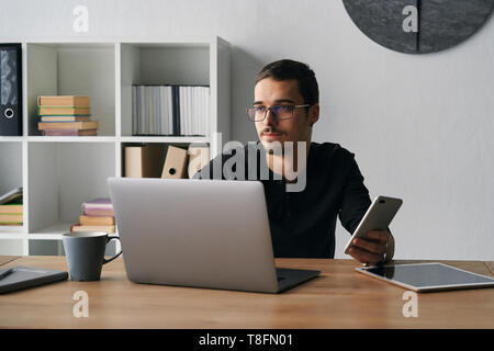 Giovane uomo che lavora con il telefono e il computer, la ricezione della chiamata di telefono, parlando con i partner Foto Stock