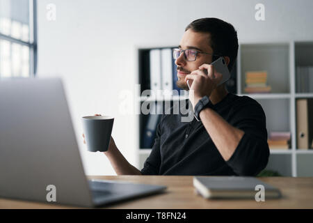 Giovane uomo che lavora con il telefono e il computer, la ricezione della chiamata di telefono, parlando con i partner Foto Stock