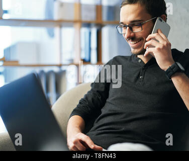 Giovane uomo che lavora con il telefono e il computer, la ricezione della chiamata di telefono, parlando con i partner Foto Stock