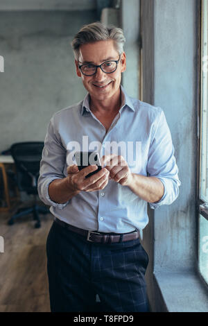 Happy businessman matura in piedi da una finestra con il suo telefono cellulare. Imprenditore maschio nel suo ufficio. Foto Stock