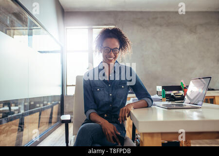 Positivo giovane imprenditrice seduto alla scrivania in ufficio. Giovane donna africana al lavoro. Foto Stock