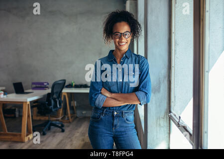 Successo femmina giovane imprenditore in piedi in ufficio con le braccia incrociate. Imprenditrice positivo in casuals in piedi vicino alla finestra in ufficio. Foto Stock