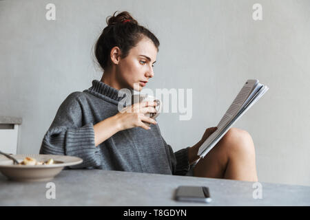 Bella giovane donna avente una sana colazione seduti su una cucina, rivista di lettura Foto Stock