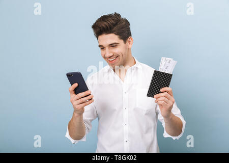 Ritratto di un allegro bel giovane uomo in piedi isolato su sfondo blu, tenendo passaporto con biglietti di volo, utilizzando il telefono cellulare Foto Stock
