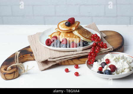 Sulla tavola è una tavola di legno con pancake con frutti di bosco freschi di ribes, mirtillo e lampone e accanto ad una tazza di fiocchi di formaggio Foto Stock