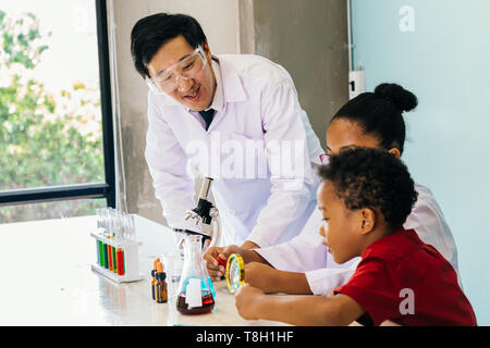 Giovane scienziato asiatico tenendo il tubo di vetro pallone e insegnamento due americano africano bambini misti in laboratorio di chimica esperimento in classe di scienze Foto Stock