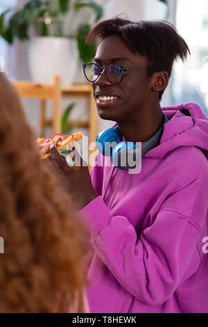 Allegro con la pelle scura adolescente di mangiare la pizza e parlando a un amico Foto Stock