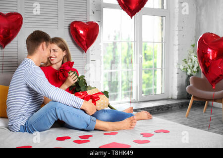 Felice coppia giovane con rose rosse e confezione regalo seduta sul letto di casa Foto Stock