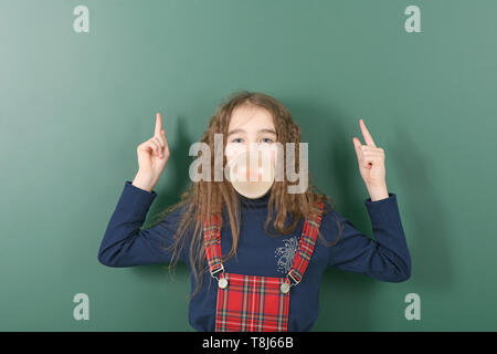 Schoolgirl vicino a green school board. Giovane ragazza giocoso mostra un dito verso l'alto e booble chewing gum. Foto ad alta risoluzione. Foto Stock