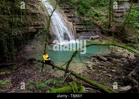 Donna in giacca gialla seduti su un ramo dalla bellissima piscina naturale, una cascata di un ruscello che scorre attraverso una folta foresta verde, Pasjak Slovenia Foto Stock