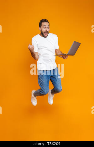 A piena lunghezza Ritratto di uomo felice 30s in t-shirt bianco azienda notebook argento isolato su sfondo giallo Foto Stock
