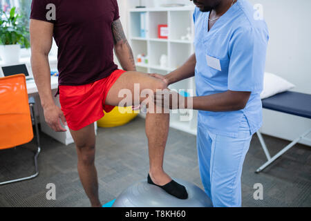 Sportivo in pantaloncini rossi mettendo gamba sulla sfera di fir in piedi vicino al terapista Foto Stock