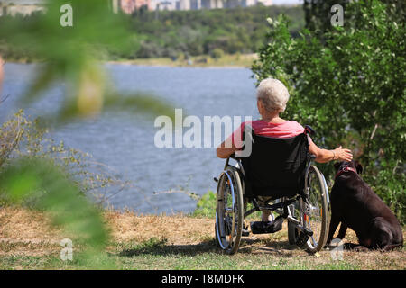 Senior donna in sedia a rotelle e il suo cane vicino al fiume Foto Stock