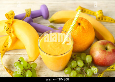 Bicchiere di gustoso frullato giallo con frutti, pesi e nastro di misurazione su un tavolo di legno. Concetto di dieta Foto Stock