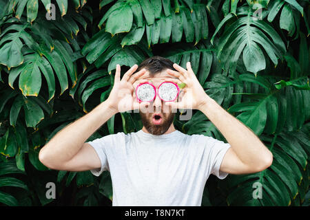 Giovane uomo barbuto holding fette di Pitaya dragon frutto di fronte ai suoi occhi, sorpreso. Foglie tropicali sfondo, copia dello spazio. Foto Stock