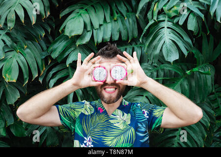 Giovane uomo barbuto holding fette di Pitaya dragon frutto di fronte ai suoi occhi, ridendo. Foglie tropicali sfondo, copia dello spazio. Foto Stock