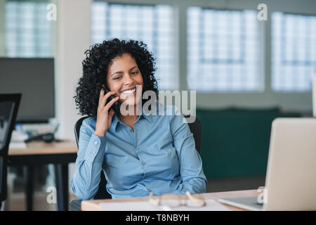 Imprenditrice sorridente seduto alla sua scrivania a parlare su un telefono cellulare Foto Stock
