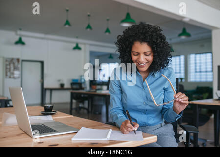 Sorridente imprenditrice seduta al lavoro di redazione di idee in un notebook Foto Stock