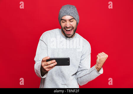 Immagine dell uomo unshaved 30s tenendo lo smartphone e la riproduzione di video giochi isolate su sfondo rosso Foto Stock