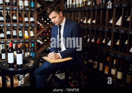 Malinconici uomo sommelier con gambe incrociate la scrittura nel blocco note somethings presso il bar. close up vista laterale foto. copia spazio. lavoro, professione Foto Stock