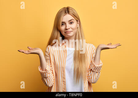 Perplesso esitante ragazza con lunghi capelli biondi vestita casualmente, alza le spalle spalle come il doesn t conosce risposta, di essere o non essere.isolato sfondo giallo Foto Stock