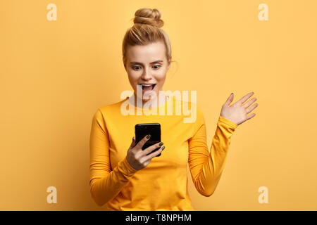 Scioccato giovane ragazza emotiva guardando il telefono cellulare isolato su sfondo giallo. close up foto. La donna ha leggere informazioni interessanti forma il gadget. Foto Stock