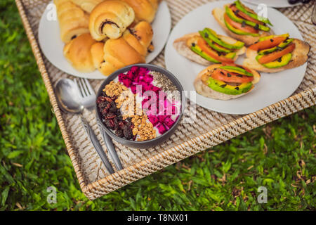 La colazione su un vassoio con frutta, panini, sandwich di avocado, smoothie ciotola in piedi sull'erba Foto Stock