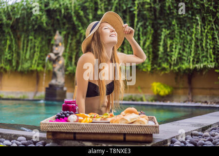 Giovane donna di mangiare la colazione su un vassoio con frutta, panini, sandwich di avocado, smoothie bowl dal pool. Estate una sana dieta vegana, prima colazione. Gustoso Foto Stock