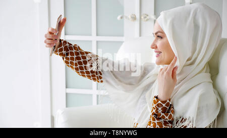 Ritratto di giovane donna musulmana in bianco hijab e abiti tradizionali è rendere selfie sullo smartphone seduto sul divano nei moderni interni bianchi, vista laterale. Foto Stock