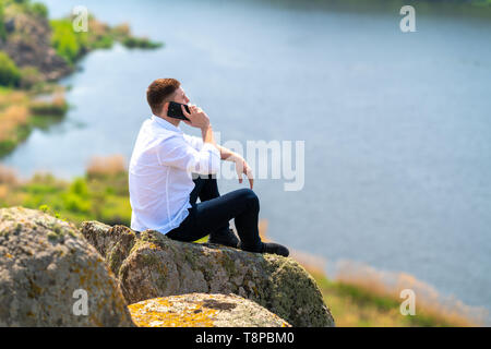Giovane uomo seduto su una roccia che si affaccia su un ampio fiume in campagna a chattare su un telefono cellulare al sole Foto Stock